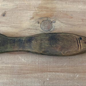 May include: A handcrafted wooden fish sculpture with a dark brown finish and carved details. The fish has a tail fin, a carved mouth, and vertical lines representing gills. The sculpture is displayed on a wooden surface.
