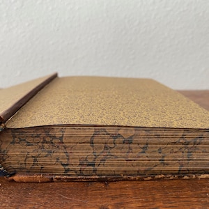 May include: A partially open antique book with a gold-toned endpaper featuring a fine dark brown pattern. The page edges display a decorative marbled design in shades of brown, black, and tan, with a worn brown leather spine and cover edges.
