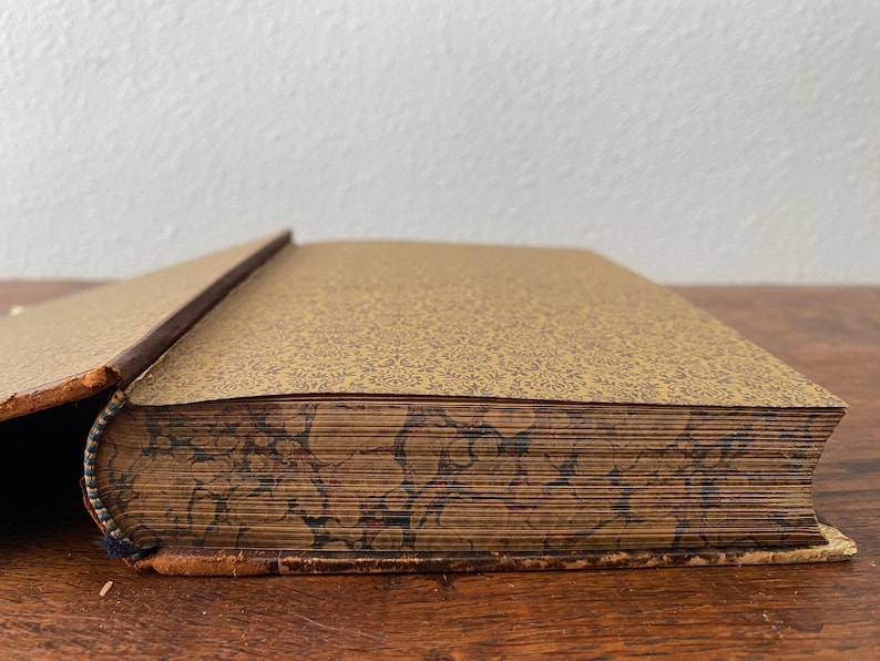 May include: A partially open antique book with a gold-toned endpaper featuring a fine dark brown pattern. The page edges display a decorative marbled design in shades of brown, black, and tan, with a worn brown leather spine and cover edges.