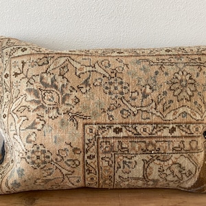 May include: A rectangular throw pillow featuring a woven textile cover with a faded, intricate floral and geometric pattern in shades of tan, beige, brown, and muted blue. The design includes a prominent border and a distressed, aged appearance.