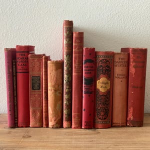 May include: A collection of ten vintage hardcover books with red and orange spines standing upright. The books feature various titles including The Scarab Murder Case, The Daffodil Mystery, and Geflügelte Worte, with decorative gold and black lettering.