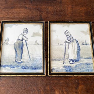 May include: Two framed rectangular wall art pieces featuring blue and white illustrations of figures in a rural landscape. The left image shows a person with a walking stick near a boat, while the right image depicts a person with a staff near a windmill.
