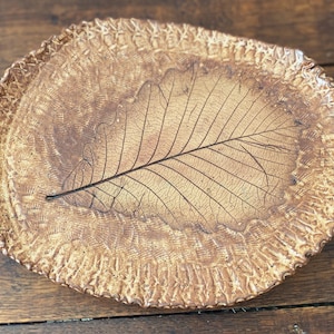 May include: An oval ceramic tray with a golden-brown glaze featuring a prominent embossed leaf imprint in the center. The surface has a textured, uneven finish with a decorative, scalloped, and ridged border surrounding the central leaf design.
