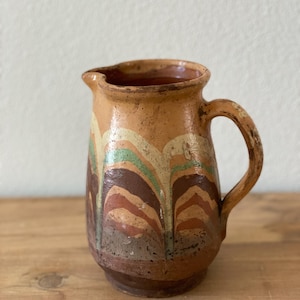 May include: A ceramic pitcher with a handle and spout. The pitcher is painted with a pattern of vertical, wavy lines in shades of brown, orange, green, and cream against a tan background. The pitcher sits on a wooden surface.