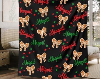 May include: Black blanket with a repeating pattern of gingerbread bow tie cookies, red and green stars, and the name "Abigail" in red and green script. The blanket is draped over a bed.