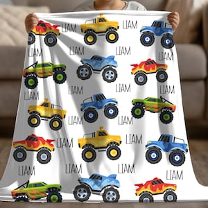 May include: A white baby blanket with a repeating pattern of colorful monster trucks. Each truck has the name "Liam" printed on it.