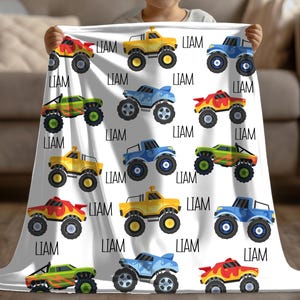 May include: A white fleece blanket with a repeating pattern of monster trucks in various colors, including yellow, blue, green, and red. The name "LIAM" is repeated throughout the blanket.