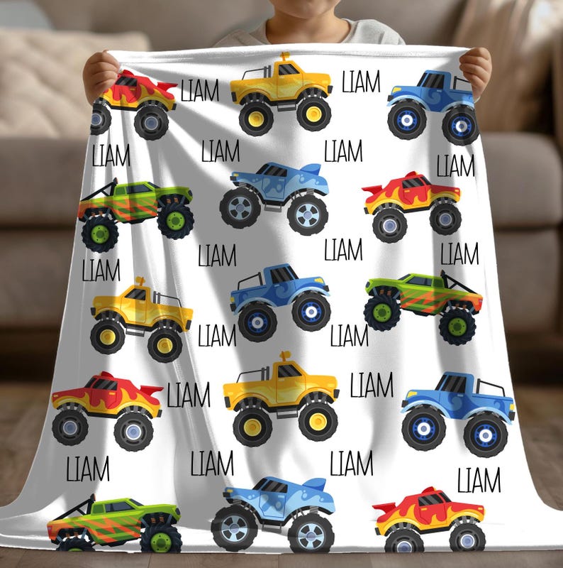 May include: A white fleece blanket with a repeating pattern of monster trucks in various colors, including yellow, blue, green, and red. The name "LIAM" is repeated throughout the blanket.