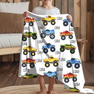 May include: A white fleece blanket with a repeating pattern of colorful monster trucks. The trucks are yellow, blue, green, and red. The name "LIAM" is printed on the blanket.