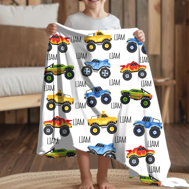 May include: A white fleece blanket with a repeating pattern of colorful monster trucks. The trucks are yellow, blue, green, and red. The name "LIAM" is printed on the blanket.