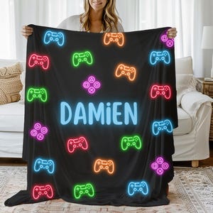 May include: Black fleece blanket with a neon green, blue, pink, orange, and yellow gamepad pattern. The blanket has the name "Damien" in a light blue font.