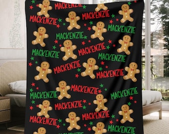 May include: Black blanket featuring a repeating pattern of gingerbread men, red and green stars, and the name "Mackenzie" in a festive font. The blanket is a cozy holiday-themed item.
