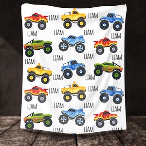 May include: A white baby blanket with a repeating pattern of colorful monster trucks. Each truck has a different color scheme and the name "LIAM" is printed on the blanket.