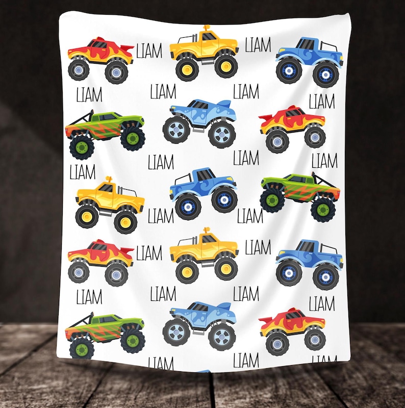 May include: A white baby blanket with a repeating pattern of colorful monster trucks. Each truck has a different color scheme and the name "LIAM" is printed on the blanket.