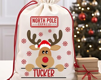 May include: A white canvas Santa sack with red drawstrings. The sack features a cartoon reindeer with a red nose and a Santa hat. The text "NORTH POLE EXPRESS" is printed in red above the reindeer. The reindeer is peeking over a sign that says "TUCKER" in red.