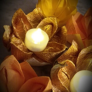 May include: Three golden paper flower candle holders with white tea lights. The candle holders are surrounded by other paper flowers in shades of orange and yellow.