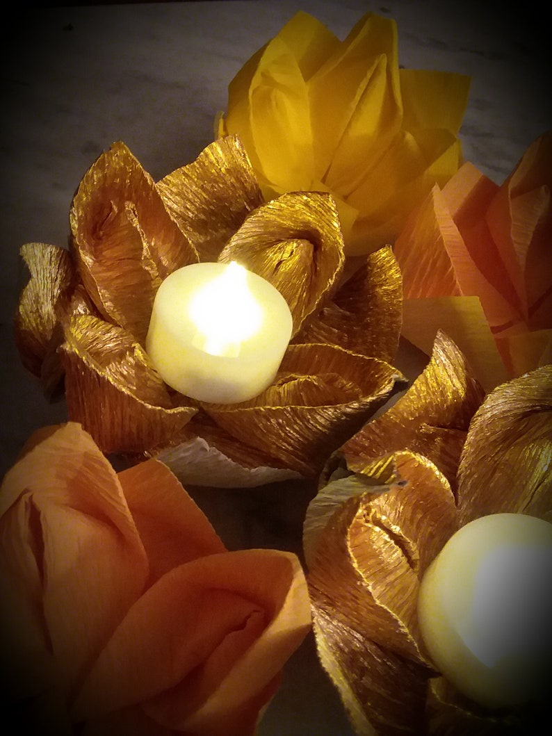May include: Three golden paper flower candle holders with white tea lights. The candle holders are surrounded by other paper flowers in shades of orange and yellow.