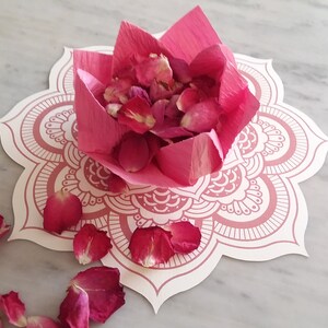 May include: Pink rose petals are arranged in a pink paper flower shape on a white mandala-patterned paper. The paper flower is on a white marble surface.