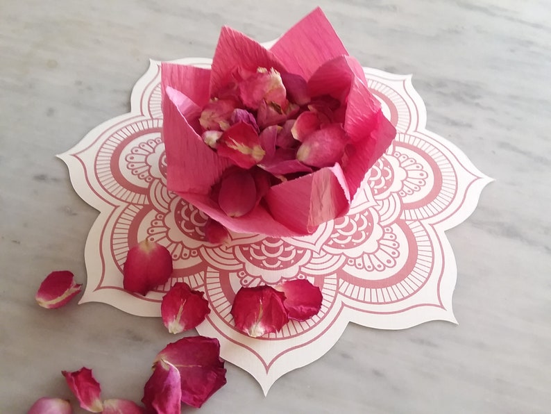 May include: Pink rose petals are arranged in a pink paper flower shape on a white mandala-patterned paper. The paper flower is on a white marble surface.