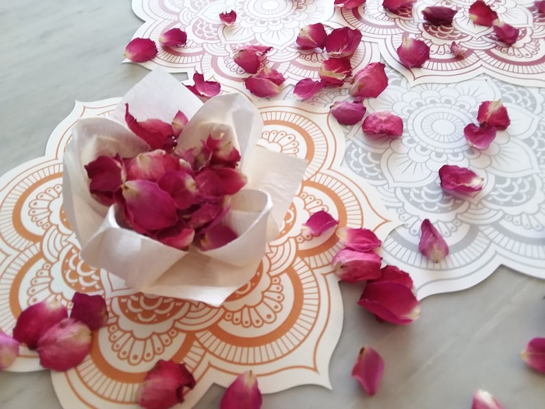 May include: A white paper cone filled with dried rose petals sits on a white and orange mandala design. The mandala is surrounded by more rose petals.