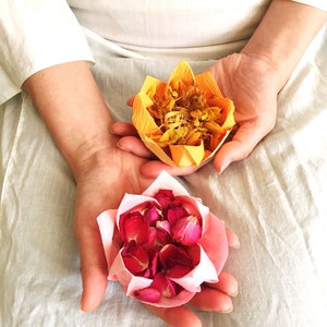 May include: Two paper flower arrangements, one yellow and one pink, are held in the hands of a person. The yellow flower is made of dried petals and the pink flower is made of dried rose petals.