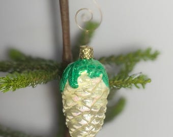 May include: A glass Christmas ornament shaped like a pinecone. The ornament has a gold-colored metal cap and a gold wire hook. The top of the pinecone is green, and the rest is a pearlescent white with glitter.