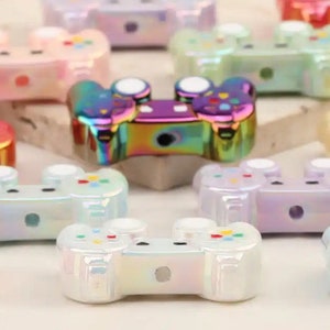 May include: A collection of iridescent game controller charms in various pastel colors, including pink, purple, and green. The controllers have a glossy finish and feature button details. These charms are ideal for jewelry making or crafting projects.