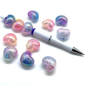 May include: Assortment of iridescent heart-shaped beads in shades of pink, purple, and blue. The beads are scattered on a white surface with a white pen.