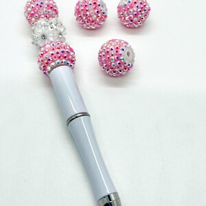 Op de afbeelding: Een witte pen met roze en witte strass steentjes die aan de bovenkant zijn bevestigd. Er zijn vier extra steentjes op de afbeelding.