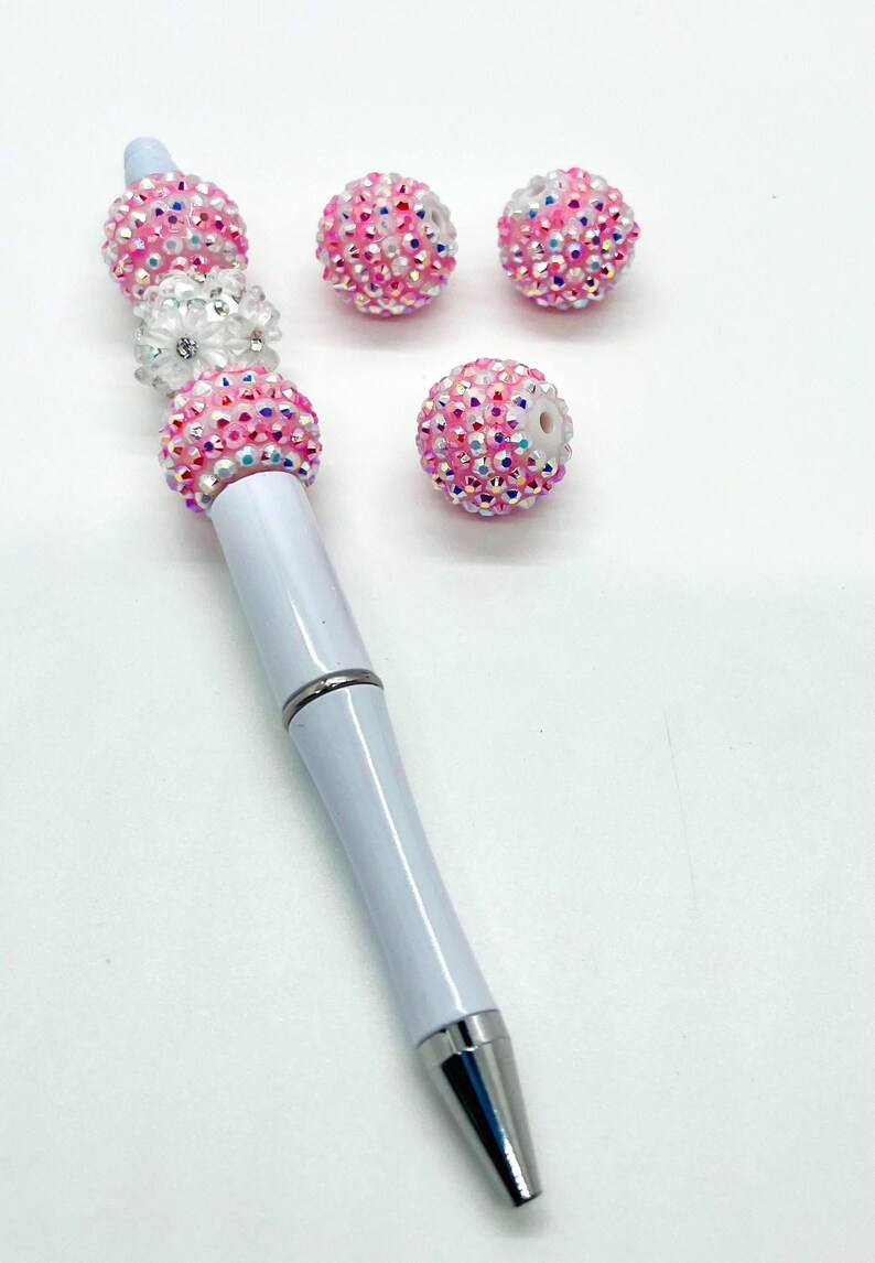 Op de afbeelding: Een witte pen met roze en witte strass steentjes die aan de bovenkant zijn bevestigd. Er zijn vier extra steentjes op de afbeelding.
