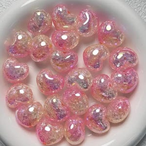 May include: A bowl filled with iridescent pink heart-shaped beads. The beads have a glittery finish and are perfect for crafting.