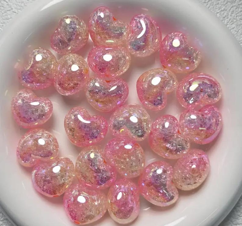 May include: A bowl filled with iridescent pink heart-shaped beads. The beads have a glittery finish and are perfect for crafting.