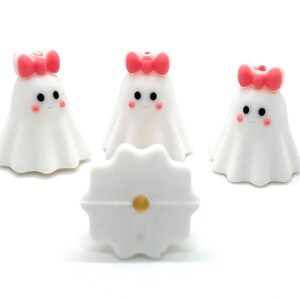 May include: Three white ghost figurines with pink bows and black eyes. The ghosts are standing on a white surface.