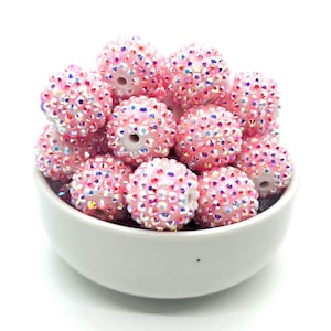 May include: A bowl filled with pink and iridescent round beads. The beads are covered in small, sparkly rhinestones.