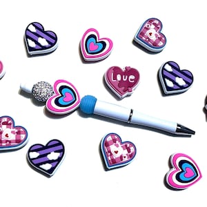 May include: A collection of heart-shaped charms in various designs and colors, including pink, purple, and blue. Some hearts feature cloud patterns, plaid, or the word "Love". A white pen-like object with a blue tip is also present.