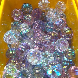 May include: A large group of iridescent glass pumpkin beads in shades of purple, blue, and clear. The beads are all approximately the same size and have a slightly textured surface.