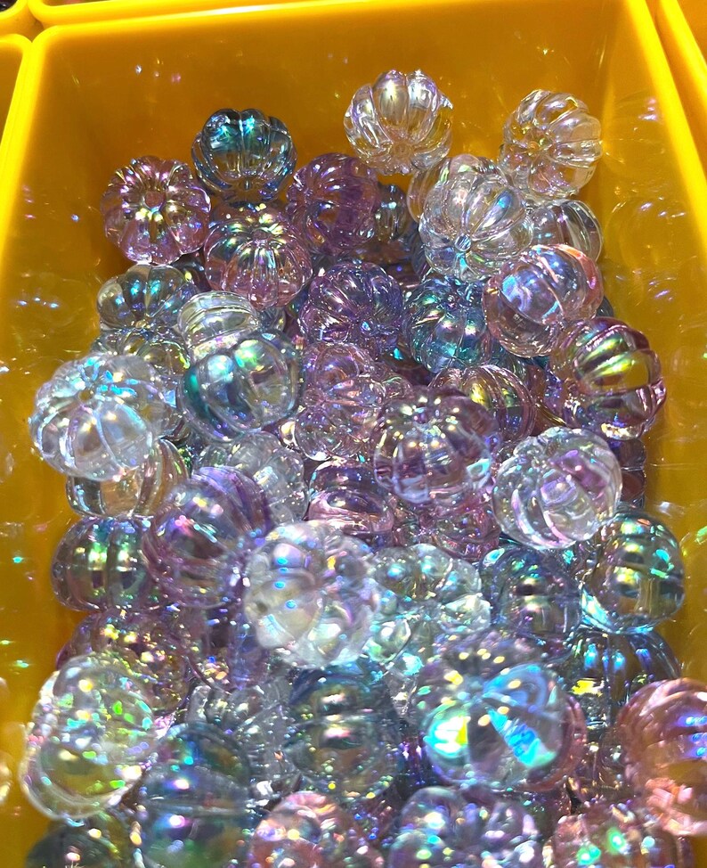 May include: A large group of iridescent glass pumpkin beads in shades of purple, blue, and clear. The beads are all approximately the same size and have a slightly textured surface.