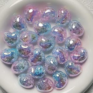 May include: A collection of iridescent heart-shaped beads in shades of pink and blue. The beads have a glittery, textured surface.