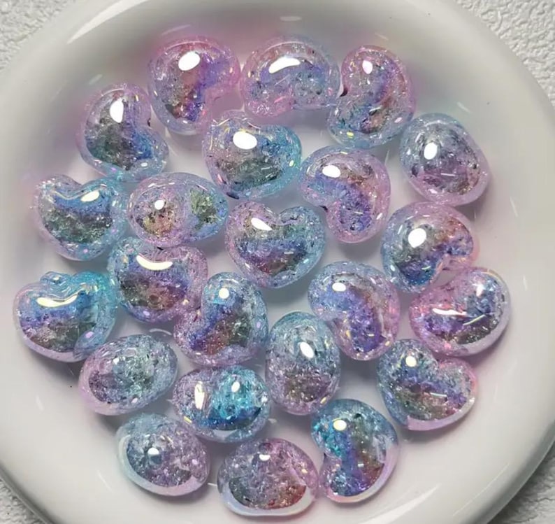 May include: A collection of iridescent heart-shaped beads in shades of pink and blue. The beads have a glittery, textured surface.