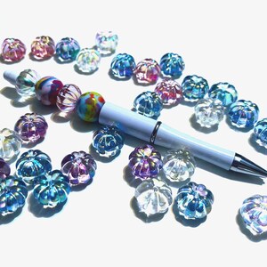 May include: A white pen with a colorful beaded design lies on a white surface surrounded by iridescent glass beads in various shades of blue, pink, and purple.