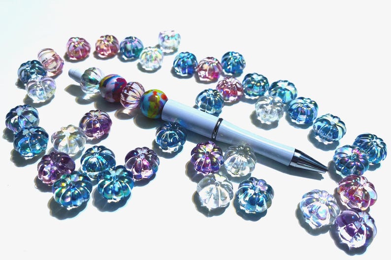 May include: A white pen with a colorful beaded design lies on a white surface surrounded by iridescent glass beads in various shades of blue, pink, and purple.