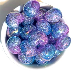 May include: A bowl filled with iridescent blue and purple heart-shaped glass beads. The beads have a textured, crackled surface and sparkle in the light.