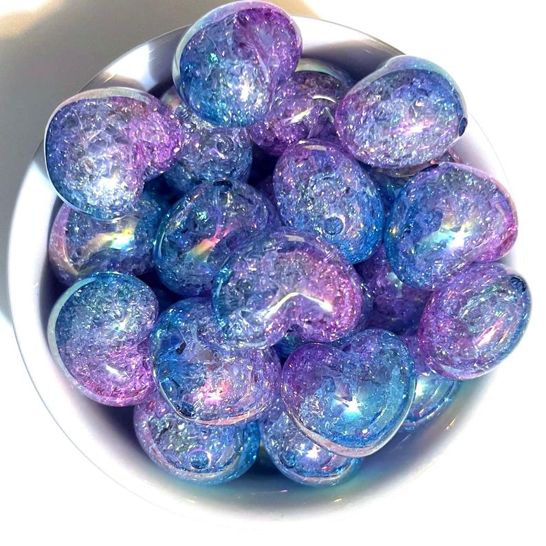 May include: A bowl filled with iridescent blue and purple heart-shaped glass beads. The beads have a textured, crackled surface and sparkle in the light.