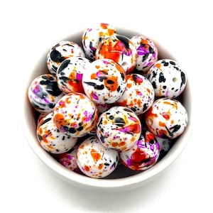 Puede incluir: Un bol de cuentas de silicona de colores con un diseño de pintura salpicada en blanco, naranja, negro y rosa.