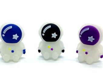 Op de afbeelding: Drie kleine astronautenbeeldjes in het wit met kleurrijke helmen. De helmen zijn blauw, zwart en paars, elk met een witte ster. De beeldjes hebben gele en paarse accenten.