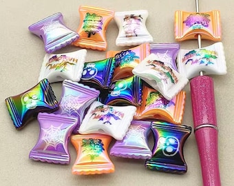 Op de afbeelding: Een verzameling kleurrijke Halloween-thema kralen in verschillende vormen en ontwerpen. De kralen tonen afbeeldingen van spinnen, schedels en de woorden "Trick or Treat". Kleuren zijn onder meer paars, oranje, wit en iriserende tinten. Er is ook een roze glitterpen aanwezig.