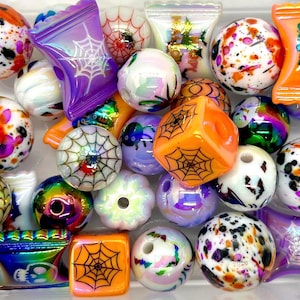 May include: A collection of Halloween-themed beads in various colors and designs, including orange, purple, white, and black. The beads feature spiderwebs, candy corn, and other spooky motifs.