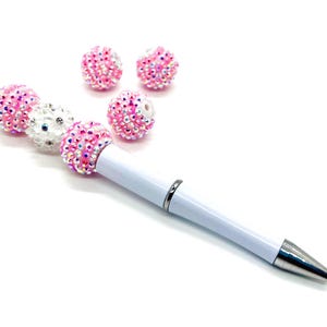 Op de afbeelding: Een witte pen met roze en witte strass steentjes die aan de pen zijn bevestigd. De pen heeft een zilveren punt en een zilveren bandje vlakbij de punt. Er zijn vier extra roze strass steentjes op de achtergrond.