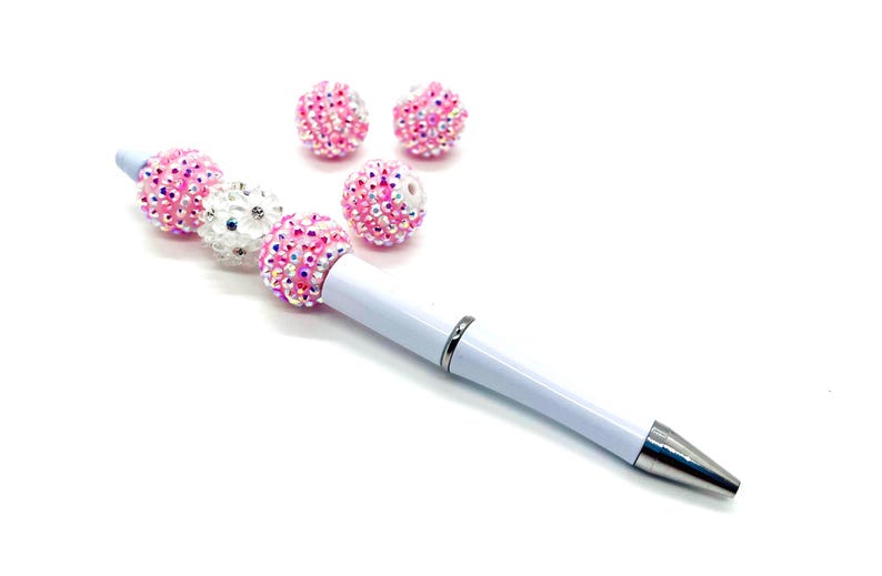 Op de afbeelding: Een witte pen met roze en witte strass steentjes die aan de pen zijn bevestigd. De pen heeft een zilveren punt en een zilveren bandje vlakbij de punt. Er zijn vier extra roze strass steentjes op de achtergrond.