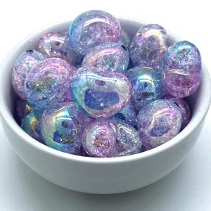 May include: A bowl filled with iridescent heart-shaped glass beads. The beads are a mix of light blue and pink with a glittery finish.